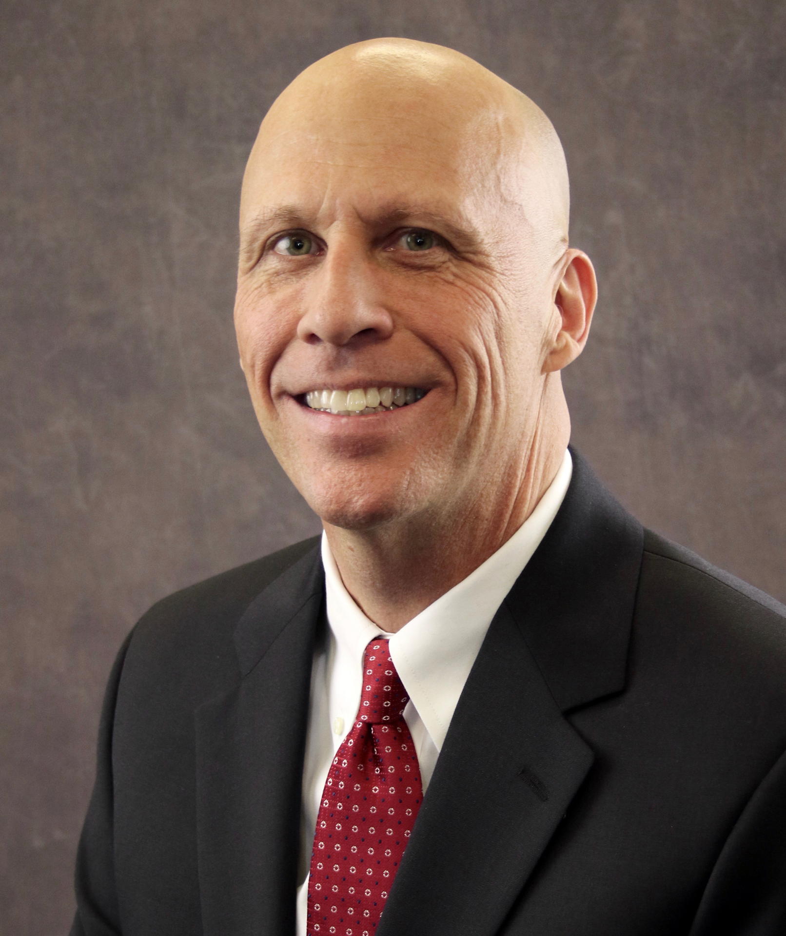Smiling bald man in a suit and red tie against a neutral background.