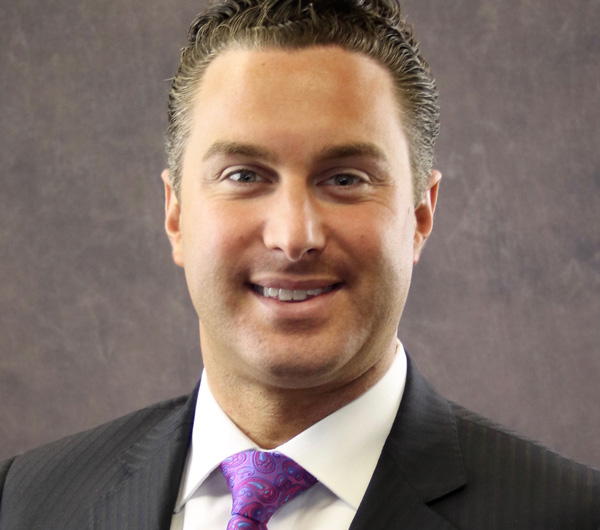 A smiling man in a suit with a purple tie against a neutral background.