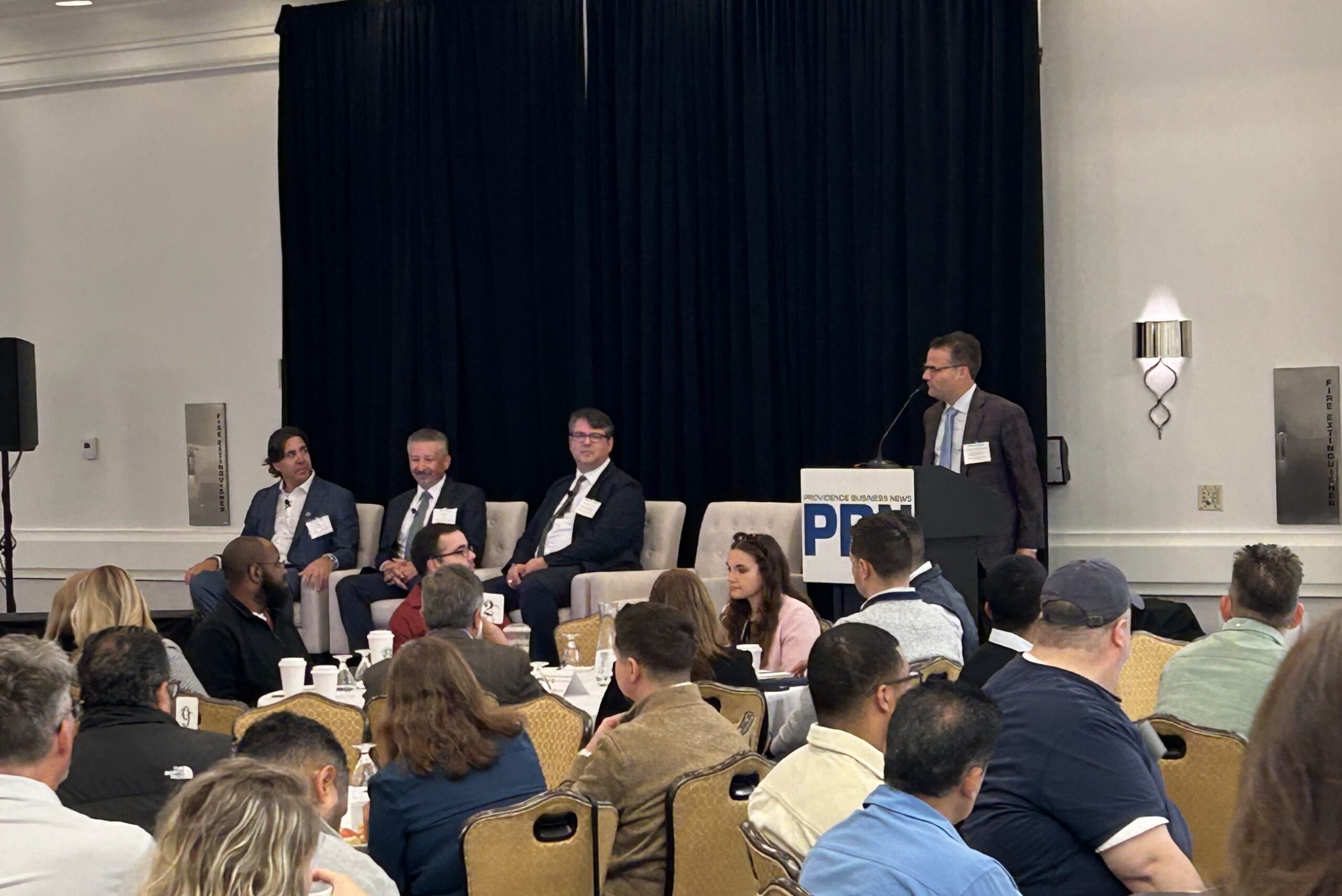 Business panel discussion with four speakers addressing attendees in a conference setting.