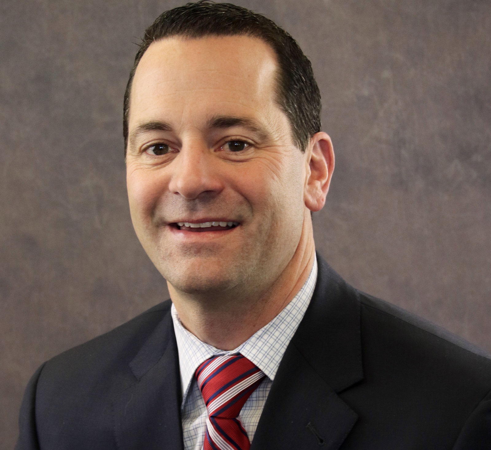 Professional man in a suit with a red striped tie, smiling against a neutral backdrop.