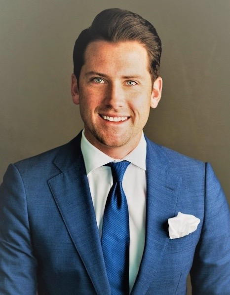 Man in blue suit with pocket square, smiling against a plain background.