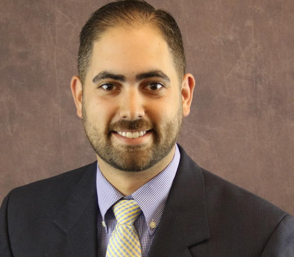 I don’t know who this is, but here is a suitable alt text: "Smiling man in business attire against a brown backdrop.