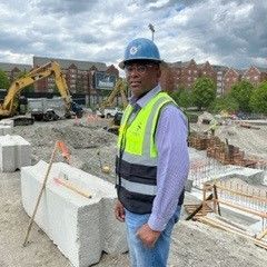 Construction site with worker in safety gear, excavator, and building foundations.