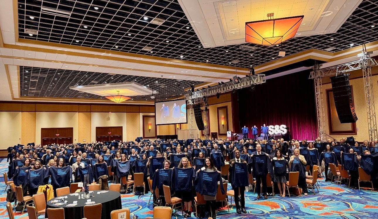 Large conference gathering with attendees holding matching shirts in a brightly lit ballroom setting.