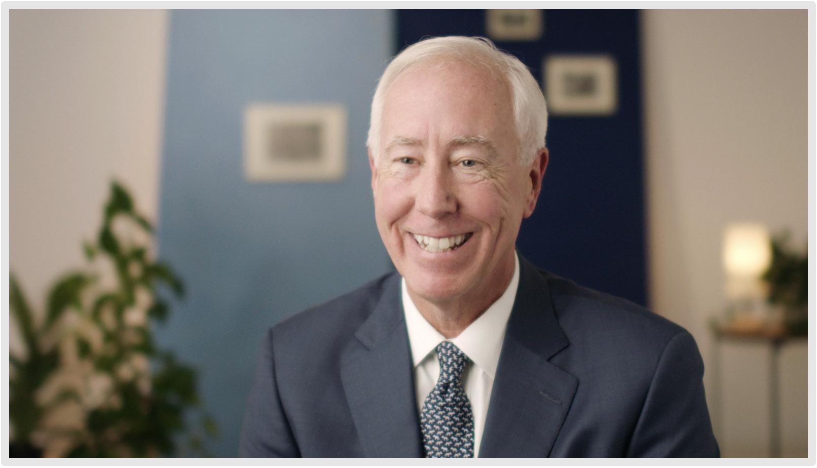 White-haired man in a suit smiling warmly in a professional setting with a blue background and plants.