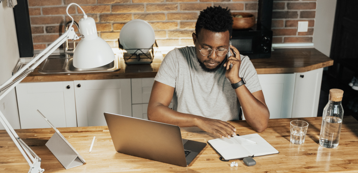 Man working from home office, using laptop and phone, with notepad and desk lamp, set in a kitchen environment.