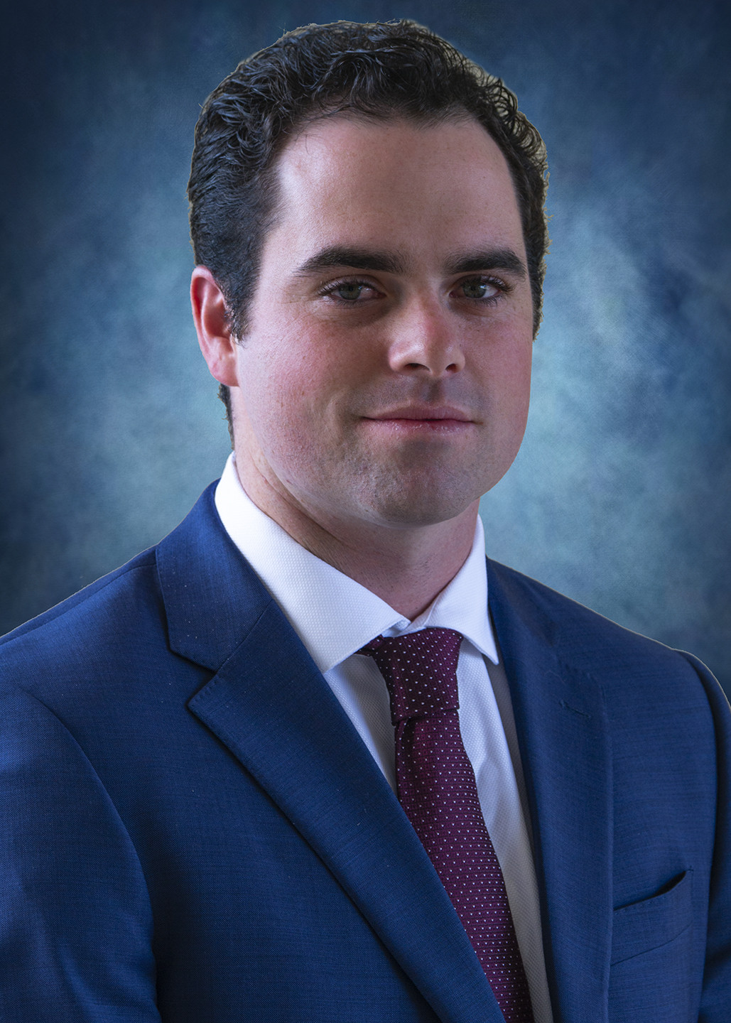 A man in a suit and tie with a neutral expression against a blue textured background.