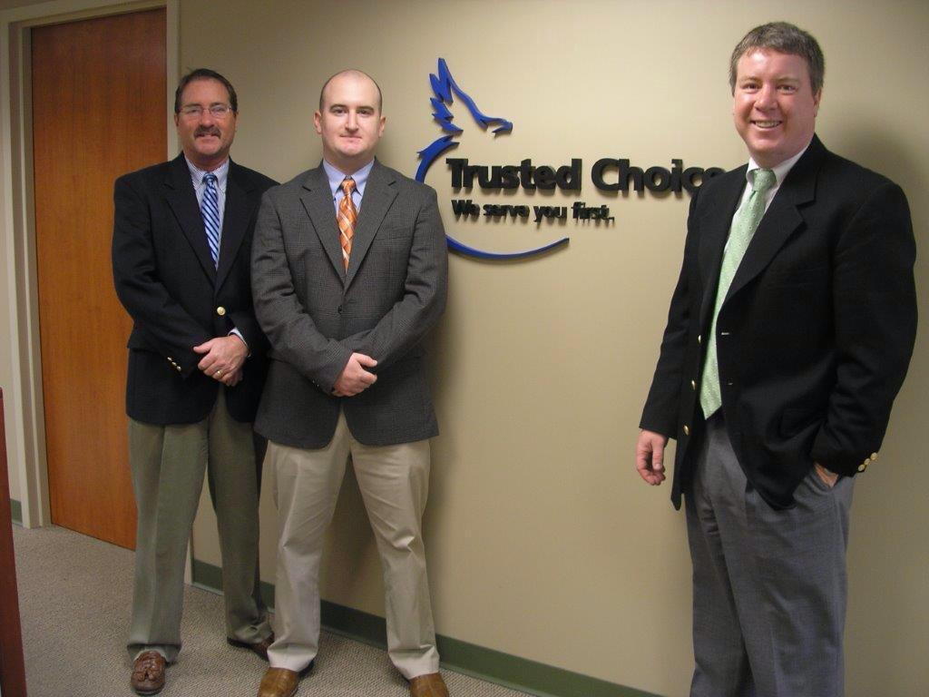 Three men standing by a "Trusted Choice" sign in an office setting, wearing professional attire.